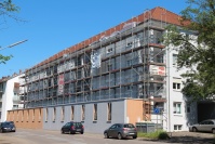 Gebäudezeile mit vier Etagen mit Baugerüst. Neue Fassadenfarbe grau, teilweise noch alte Farbe. Vorne Straße.