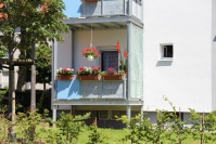 Balkon mit Blumenkästen und Hängeampel, davor Wiese und Baum.