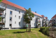 Hellgraues Wohnhaus mit neuem Dach, angebauten Balkone, 3 Etagen. Davor eine Wiese mit kleinen Bäumen.