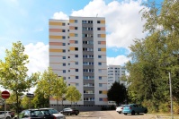 2 quadratische Hochhäuser mit 12 Etagen. Fassaden weiß. Farbakzente gelb/orange Haus vorne, hinten blau. Vorne Parkplatz mit Autos und Bäumen.