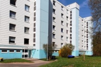 Wohnhaus 6 Etagen, Flachdach. Weiße Plattenfassade, Treppenhaustürme türkis. Davor asphaltierter Weg.