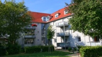Graues Wohnhaus mit 4 Etagen, angebautwe Balkone, Dach neu. DAvor Wiese und hohe Bäume am Rand.
