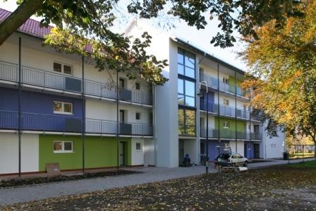 Wohnhaus mit Laubengängen, links drei Etagen, rechts vier Etagen. Vorne Blätter von einem Baum. Wohnhaus mit Laubengängen, links drei Etagen, rechts vier Etagen. Vorne Blätter von einem Baum.