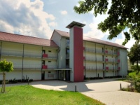 Wohnhaus mit zwei Baukörpern und Aufzugsturm davor. Links drei Etagen, rechts vier Etagen. Davor gepflasterte Fläche. 
