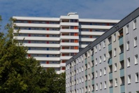 Hinten weißes Hochhaus mit Laubengängen und Aufzugsturm, vorne rechts 6geschossiges Flachdachhaus mit gelben Loggien, Fassade hellblau. Links hohe Bäume im Sommer.
