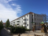Wohnhäuser mit Satteldach, graue Fassade, weiße Balkonbrüstungen. Parkplätze mit Autos, rechts Spielplatz. Wohnhäuser mit Satteldach, graue Fassade, weiße Balkonbrüstungen. Parkplätze mit Autos, rechts Spielplatz.